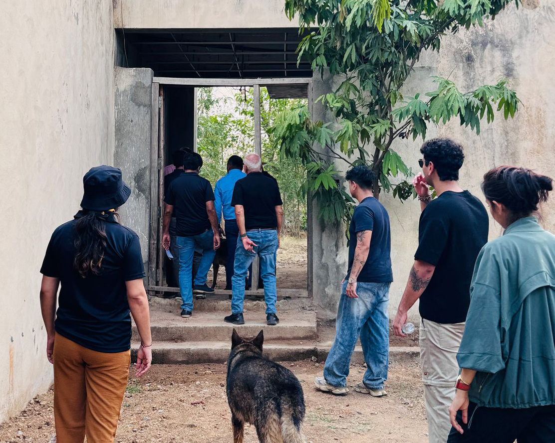 A group of people and a German Shepherd dog walking toward the entrance of a modern, minimalist concrete building surrounded by lush green trees.
