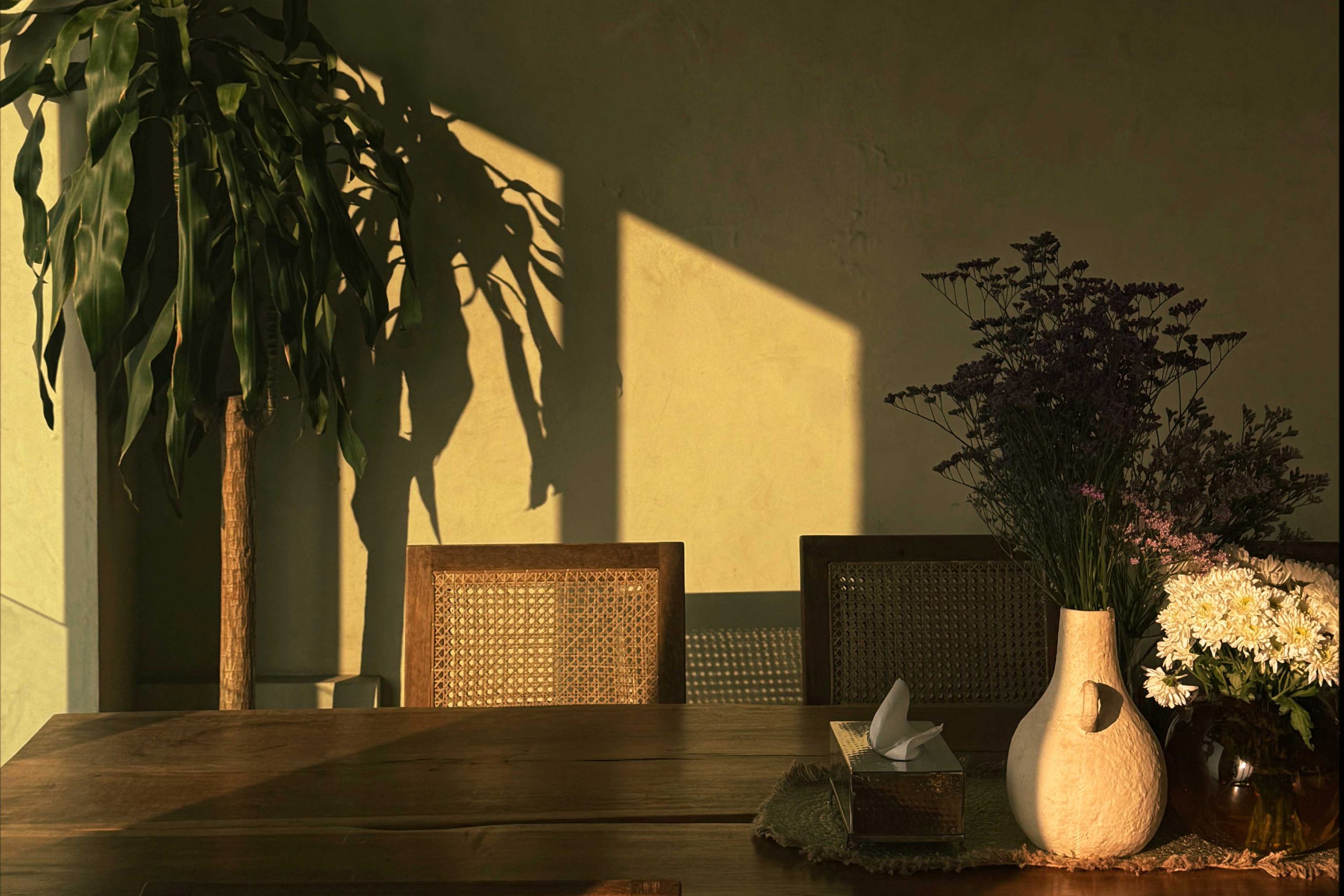 Sunlit wooden dining table with cane chairs and floral arrangement indoors.