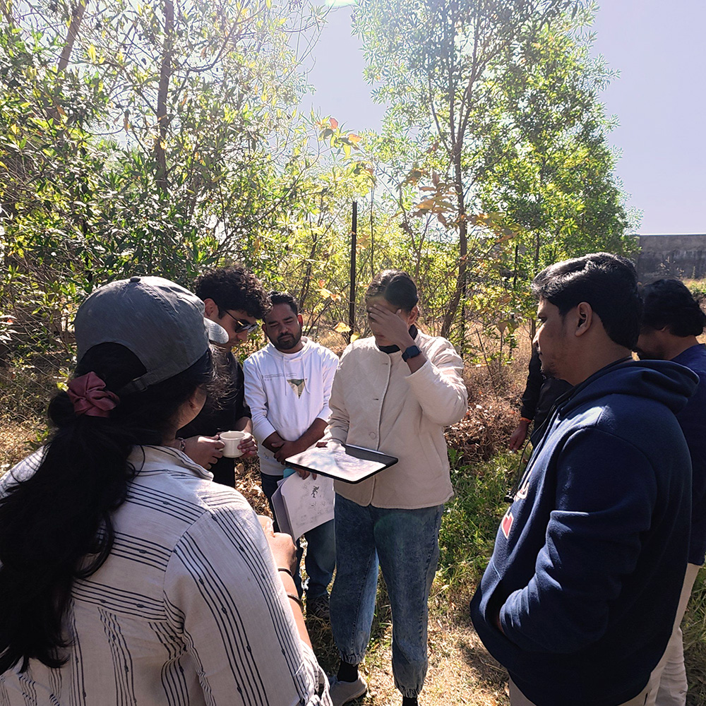 A group of people standing outdoors in a forested area, gathered closely while one person looks at a tablet.