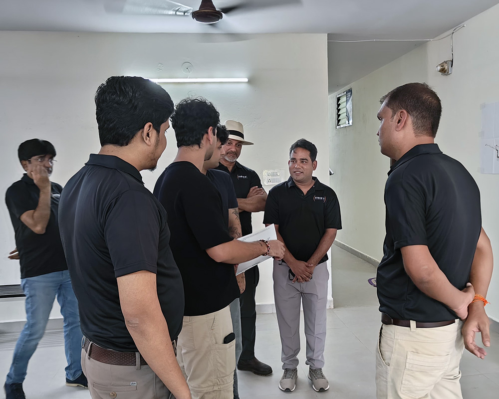 A team of professionals in black polo shirts engaged in an indoor meeting, standing in a circle and discussing documents in a brightly lit, white-walled office space.