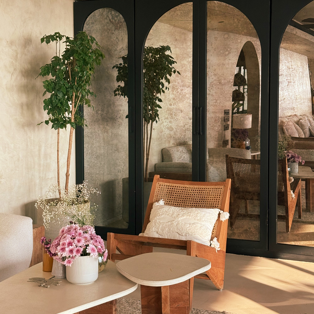 A bright sitting area with a wooden armchair, a white side table with pink flowers, and large arched mirrors in the background.
