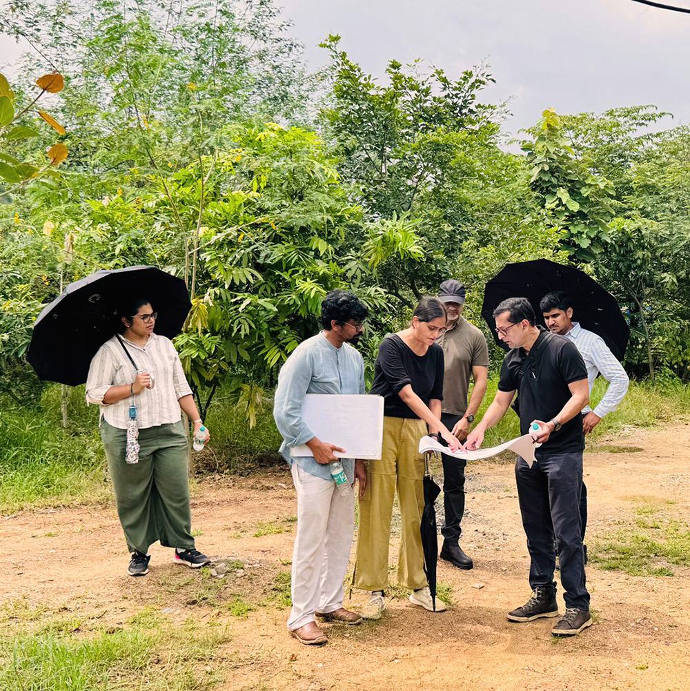 A group of people standing on a dirt path surrounded by greenery, reviewing papers together, some holding umbrellas.