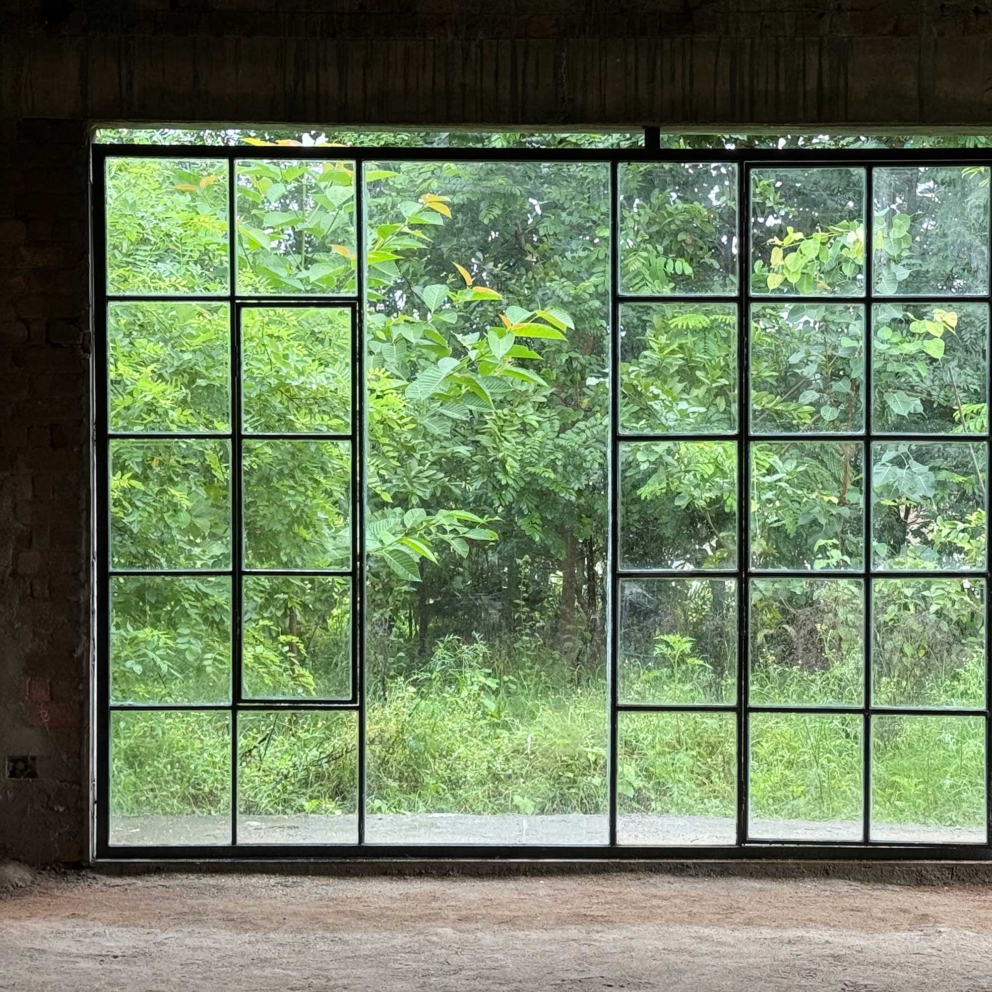Large grid-style window looking out onto dense green foliage and trees, with soft natural light entering an industrial interior space.