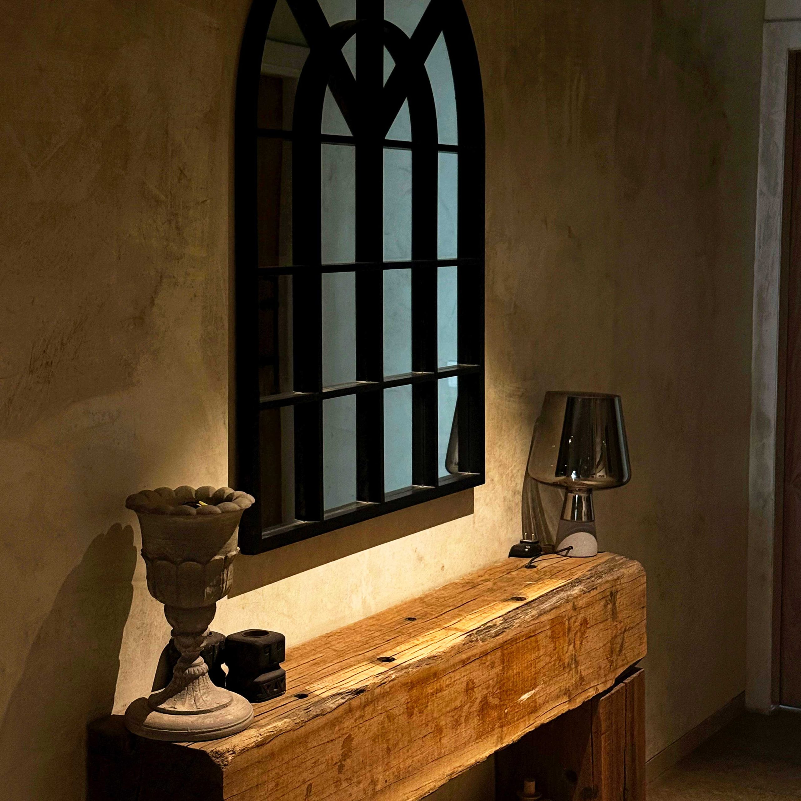 A rustic, thick-cut wooden console table in a dimly lit hallway beneath an arched window-style mirror.