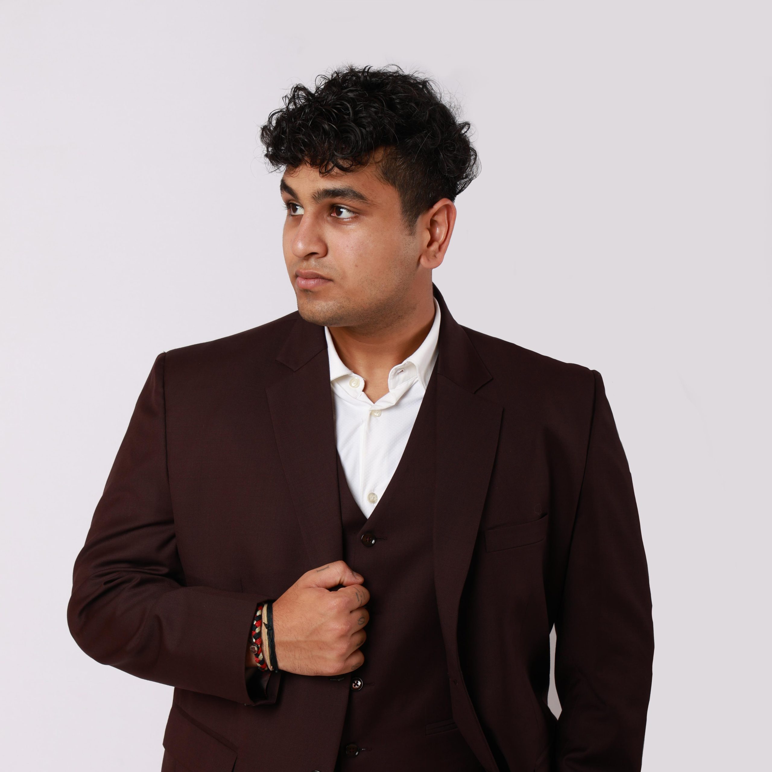 A professional studio portrait of Maurya Shah, wearing a dark brown three-piece suit and a white dress shirt, looking thoughtfully to the side against a plain white background.