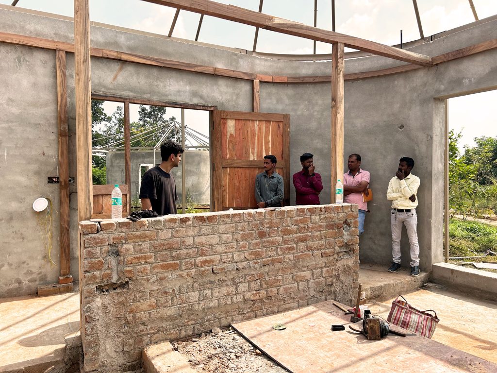 A construction site meeting where a group of men, including one of the founders, stand inside a circular building under construction with exposed brick walls and timber roof framing.